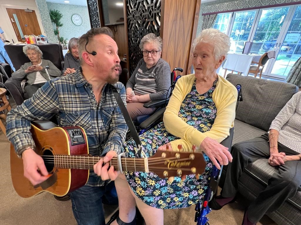 guitarist with headset microphone kneels down to sing to care home resident in chair