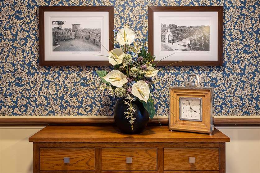 a wooden sideboard in front of blue floral wallpaper with a plant and clock